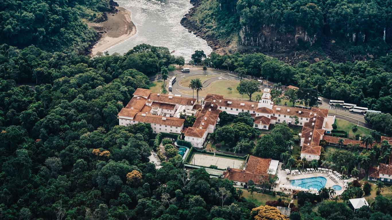 Aerial view of the Hotel das Cataratas nestled within the lush greenery of Parque Nacional do Iguaçu, with the Iguazu River and waterfall visible in the background. The hotel's red-tiled roofs and white walls stand out amidst the surrounding dense forest.