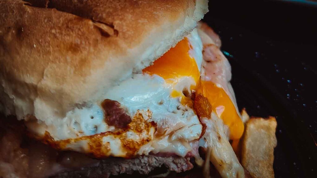 A close-up of a Chivito, Uruguay's classic sandwich, with a tender slice of steak topped with melting cheese, a perfectly cooked egg, and crispy bacon peeking out of a toasted bun.