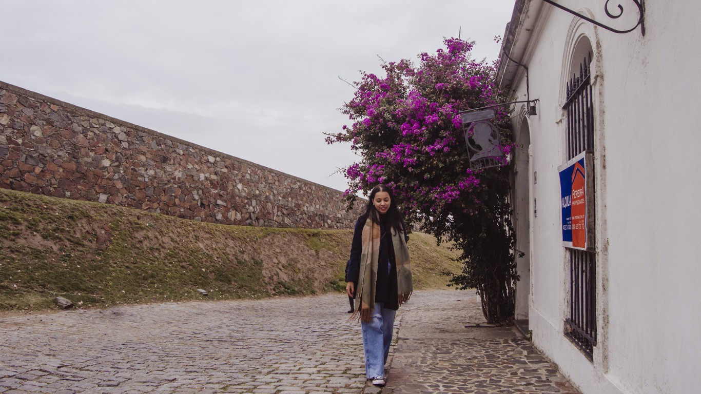 The image shows a young woman walking along a cobblestone street lined with a white building. She is dressed in casual attire with a long scarf and coat. On the right side of the image, a bright pink bougainvillea bush is in full bloom, climbing up the building's wall. There is also a sign attached to the building and a stone wall running parallel to the street in the background. The overall scene gives a serene, quaint atmosphere, possibly in a historic or scenic area.