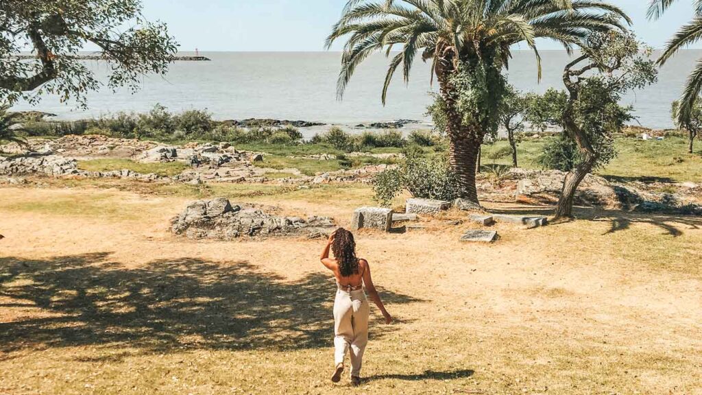 A woman in summer attire walks toward the tranquil shores of Rio de la Plata, framed by verdant trees and the warm golden sands of Colonia del Sacramento's coastline.