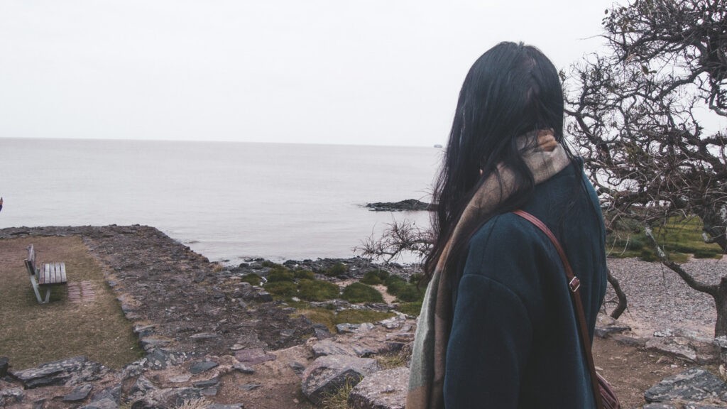 A contemplative figure gazes out over the tranquil waters of Río de la Plata from a rocky vantage point, framed by the rugged beauty of Colonia del Sacramento's coastline.