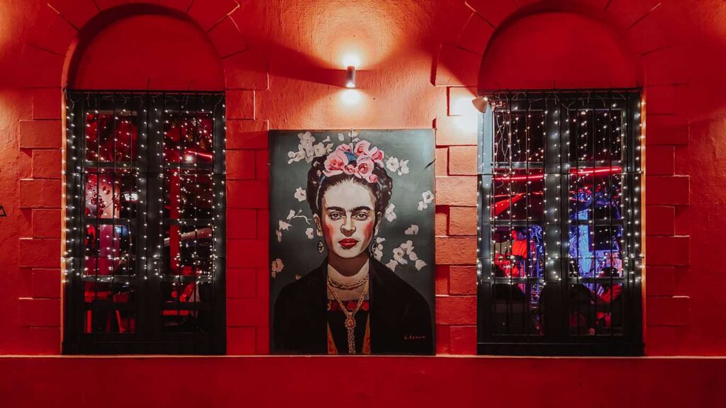 A vivid portrait of Frida Kahlo adorns a striking red wall between windows draped with twinkling lights, offering a glimpse into the cultural and artistic vibe one might find in Colonia del Sacramento.