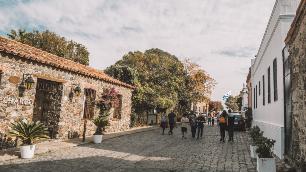 Visitors stroll through a picturesque street in Colonia del Sacramento, flanked by a stone facade with the sign "CHARCO" and blooming bougainvillea, reflecting the charming and historic atmosphere of the town.