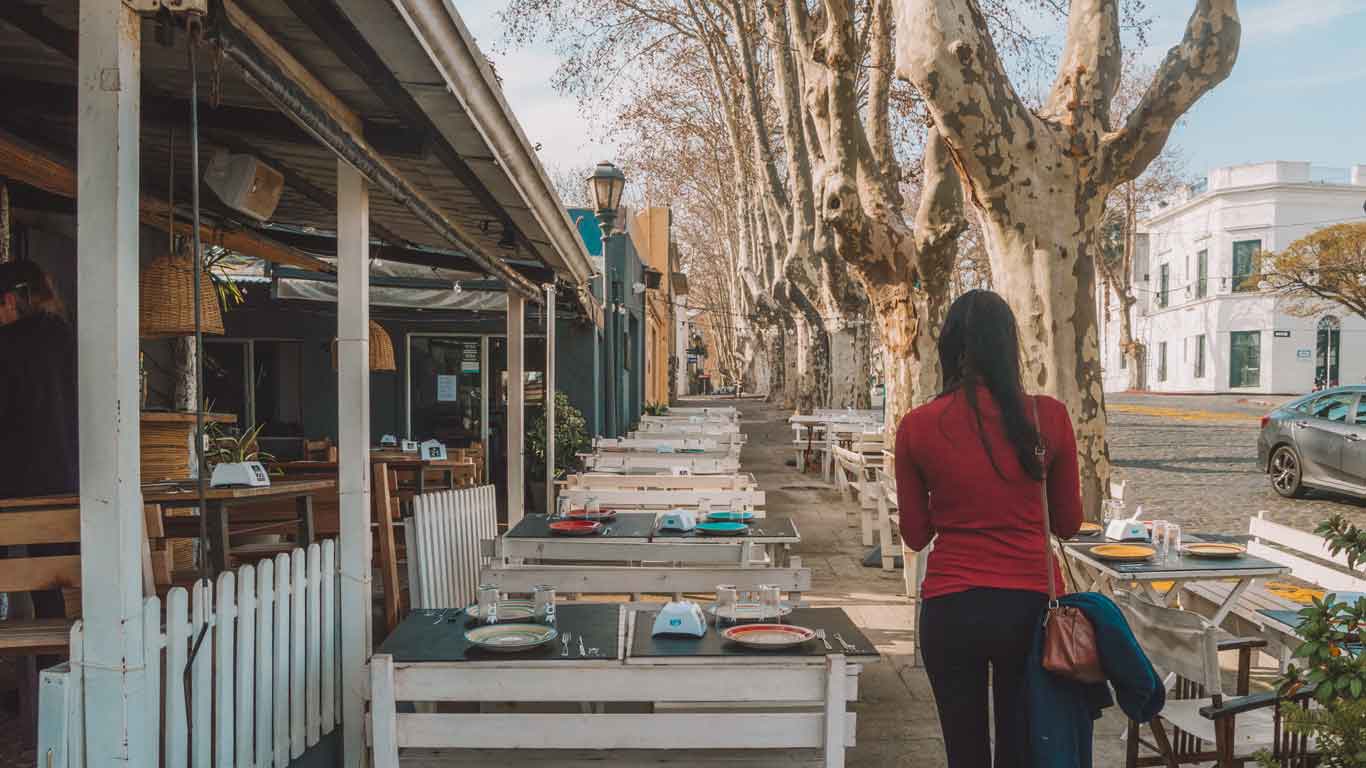 A woman in a red top walks past an inviting outdoor dining area of a restaurant in Colonia del Sacramento, lined with sycamore trees and set against the backdrop of historic architecture, epitomizing the city's cozy and welcoming atmosphere.