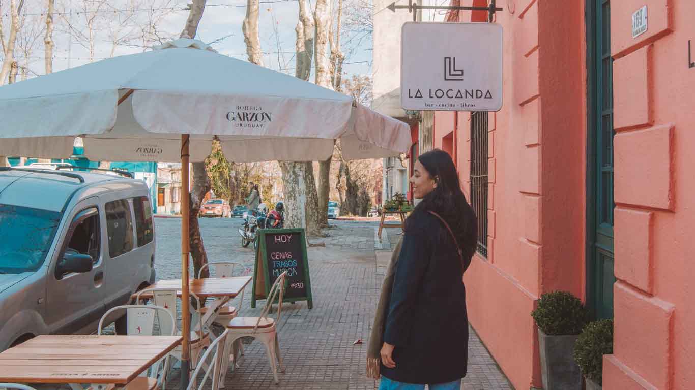 A woman contemplates where to dine at a cozy sidewalk setting outside 'La Locanda', against a backdrop of a coral-colored wall in Uruguay, evoking the inviting atmosphere of South American eateries.