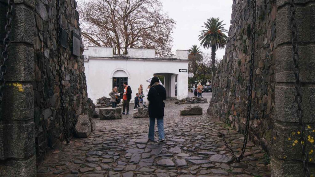 A person in a black coat stands centered between two ancient stone walls, leading to a white archway where visitors gather, under a grey sky in Colonia del Sacramento.