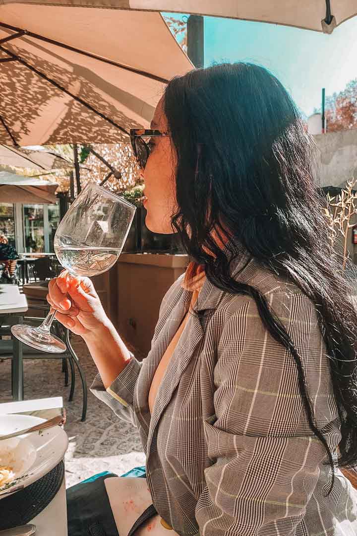 A woman enjoys a glass of white wine at an outdoor restaurant in Colonia del Sacramento, with the sunlight dappling through the umbrella, suggesting a leisurely dining experience in this charming Uruguayan city.