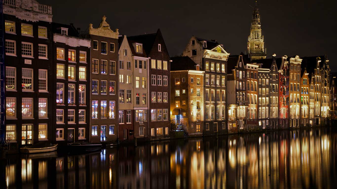 Traditional houses facing the canal in Amsterdam, illuminated at night with colorful reflections in the water. The characteristic architecture and soft lighting highlight the historical beauty of the city. In the background, a church tower rises against the night sky.