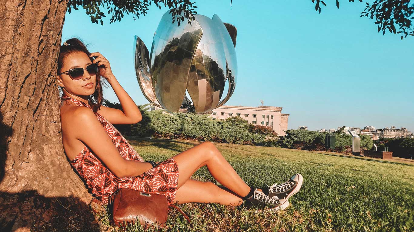 A woman sitting in the shade of a tree, wearing sunglasses and a red dress, with the famous Floralis Genérica sculpture in the background in Buenos Aires. The day is sunny, and the green landscape of the park contrasts with the light blue sky.