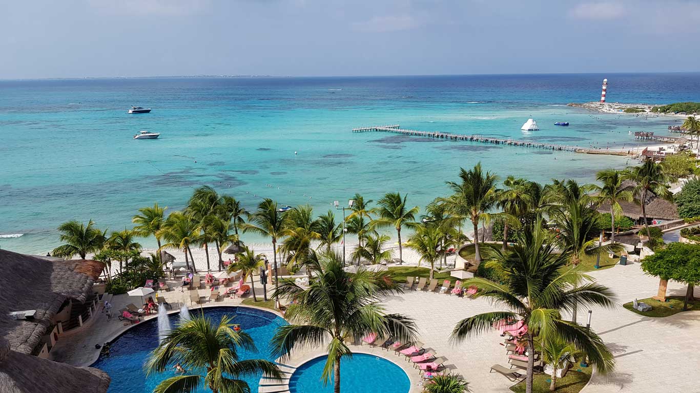 Aerial view of a paradisiacal beach in Cancún, Mexico, with crystal-clear turquoise waters, several palm trees, lounge chairs around a pool, and boats anchored in the sea. In the background, there is a pier extending into the ocean and a lighthouse at the tip of the peninsula, characterizing one of the best places to travel with friends.