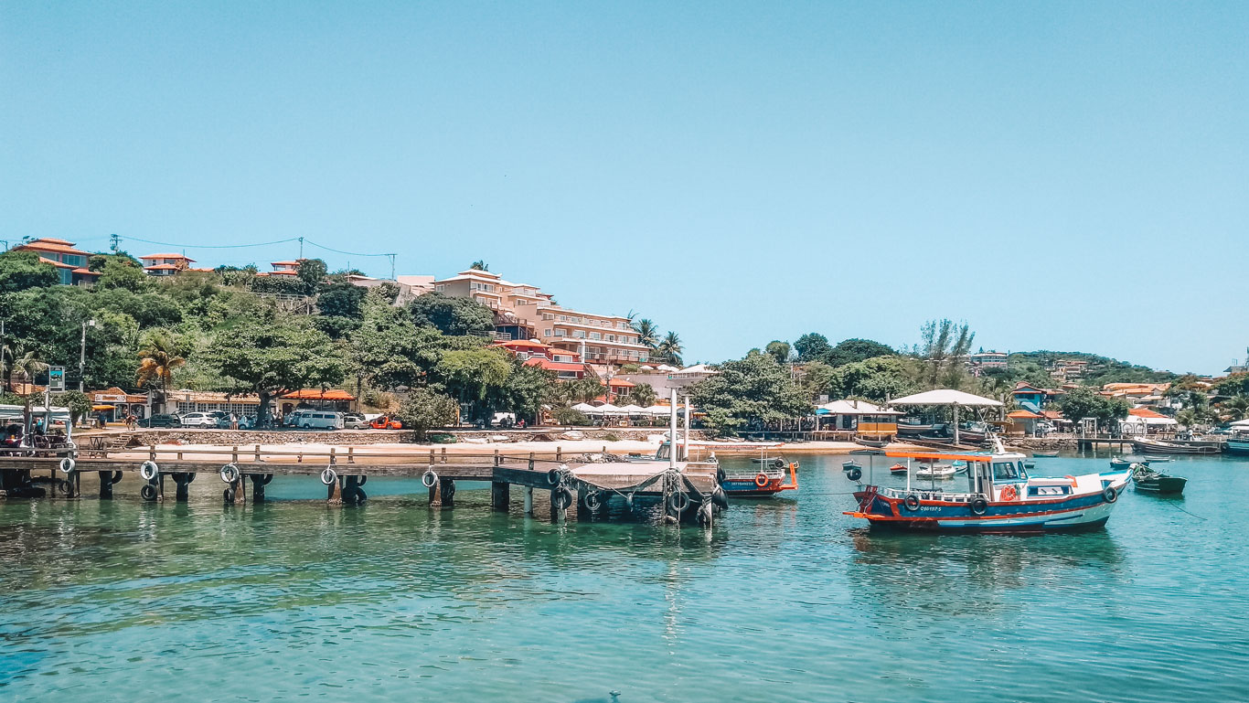 Calm waters at the harbor in Buzios, Brazil, with a wooden pier leading to colorful boats moored alongside, set against a backdrop of terracotta-roofed buildings nestled among tropical foliage under a clear blue sky.