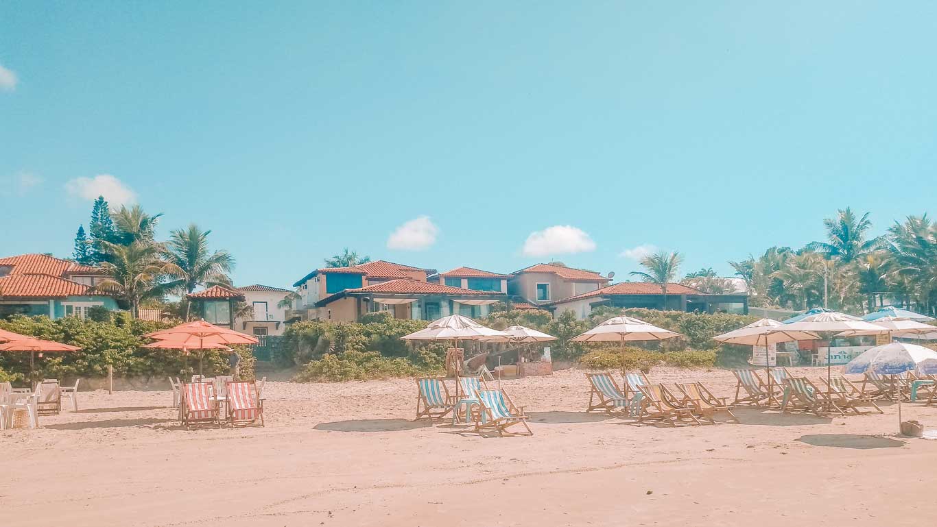 Sunny day at Geribá Beach with rows of inviting beach chairs and open umbrellas on the sand, fronting charming houses with red-tiled roofs and lush palm trees, capturing the essence of a relaxing getaway in Buzios, Brazil.