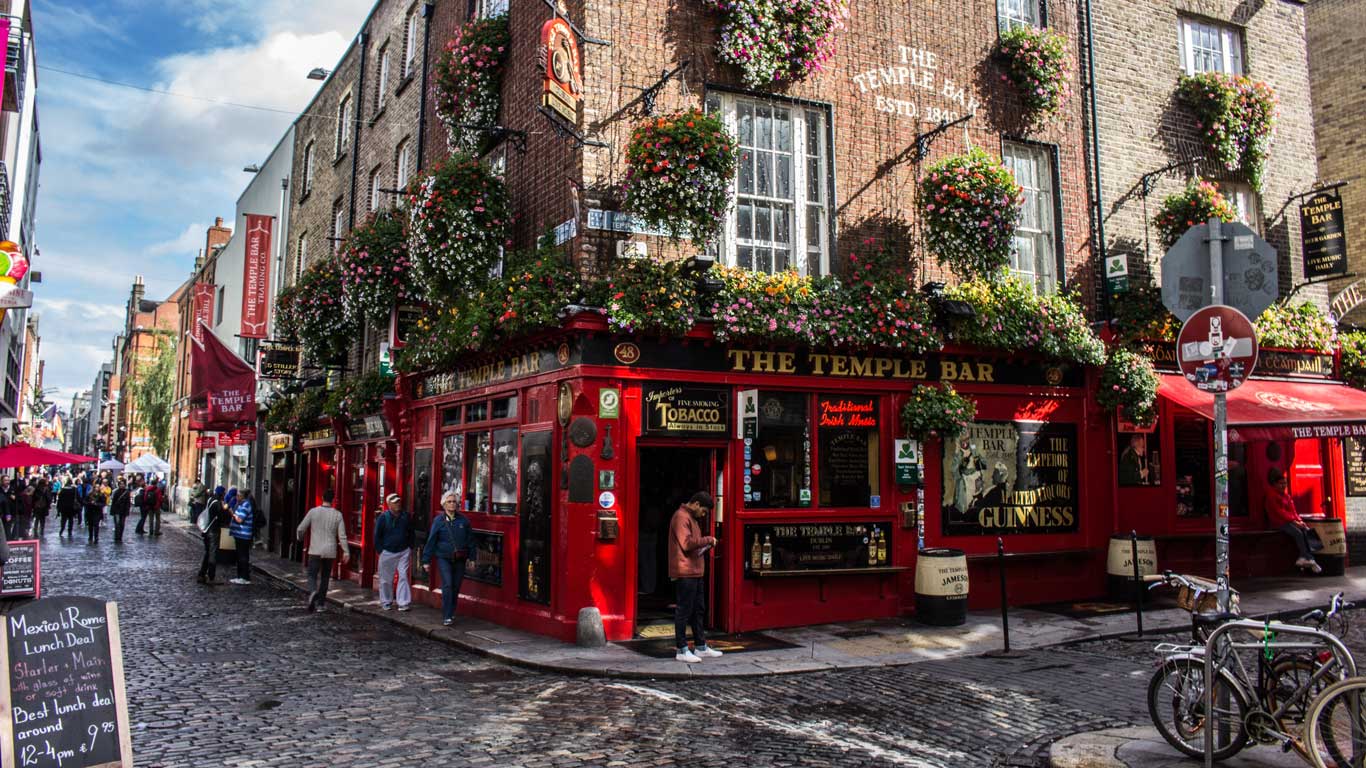 Facade of the famous pub The Temple Bar in Dublin, with its vibrant red paint and colorful flower baskets hanging from the windows. The cobblestone street around it is bustling with pedestrians exploring the historic and cultural area of the city.