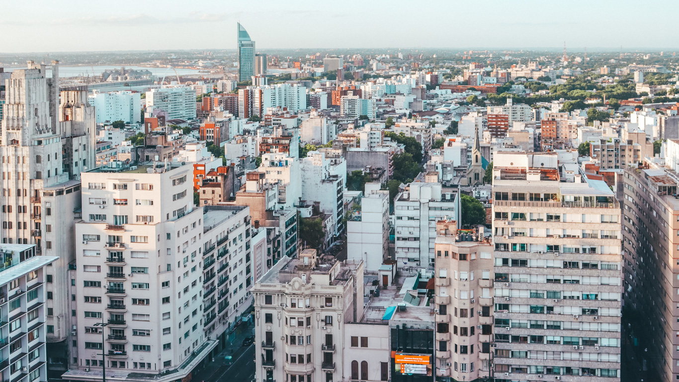 A panoramic view of Montevideo's cityscape, showcasing a mix of modern and historic buildings under a clear sky. The skyline features various high-rise structures, including the distinctive Torre de las Telecomunicaciones, with the vast expanse of the city stretching into the distance.