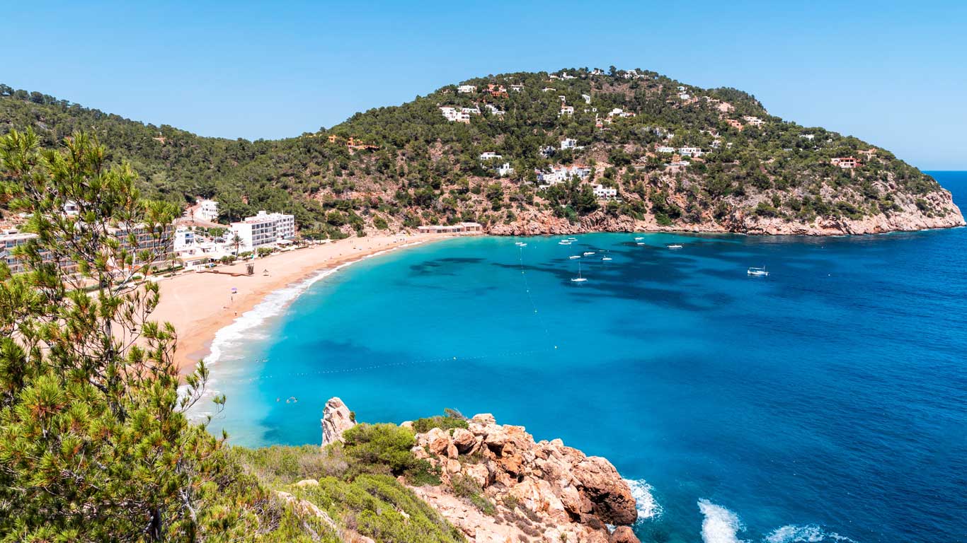 Panoramic view of a golden sandy beach in Ibiza, Spain, surrounded by green hills and crystal-clear turquoise waters. Several boats are anchored in the tranquil bay, and there are white buildings along the coast.