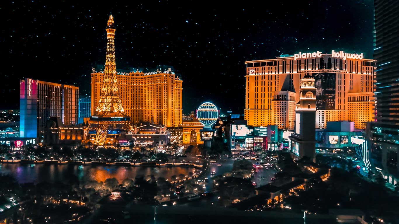 Night view of the Las Vegas Strip, featuring the Paris Las Vegas hotel and its illuminated replica of the Eiffel Tower. The Planet Hollywood hotel and other iconic casinos shine in the background under a starry sky, showcasing the vibrant nightlife of the city.