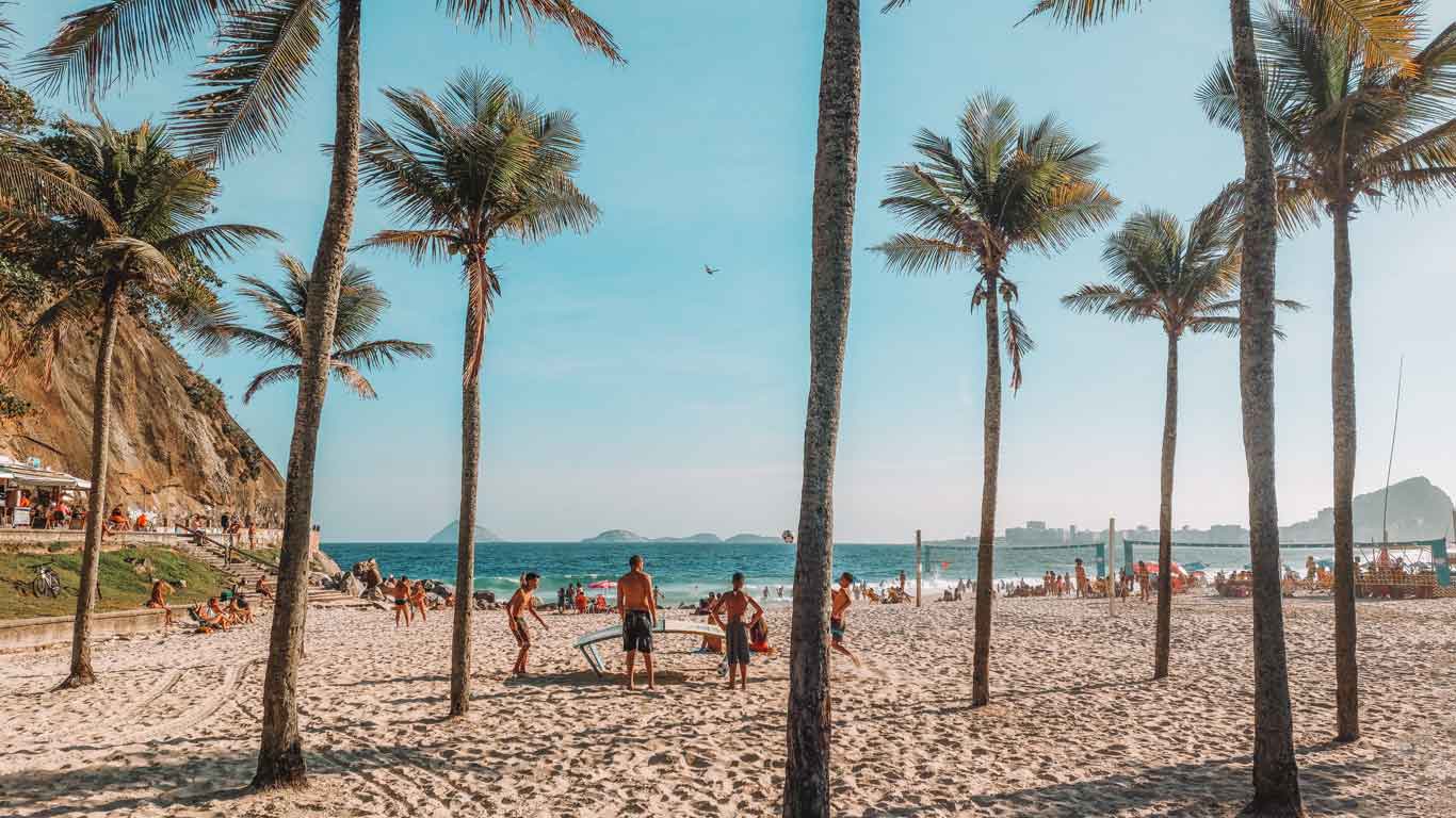 Leme Beach in Rio de Janeiro, with people enjoying the sunny day among palm trees and light sand. Groups are playing volleyball and paddleball, while others relax in the shade. The blue sea and clear sky complement the vibrant and lively scene.