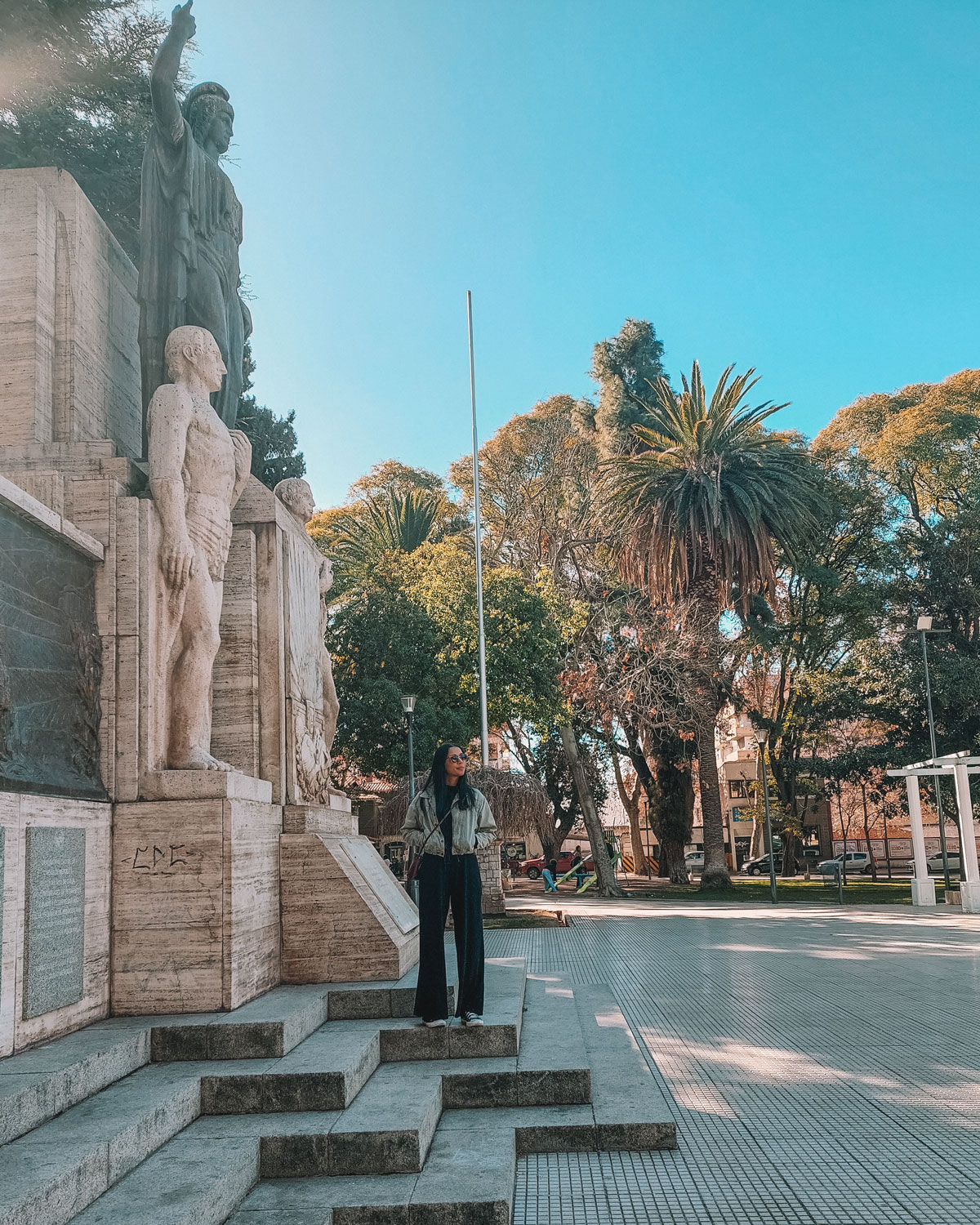 A person stands at the base of a grand statue under a clear blue sky in Mendoza, with vibrant green trees in the background, providing a serene setting in this picturesque Argentinian city.