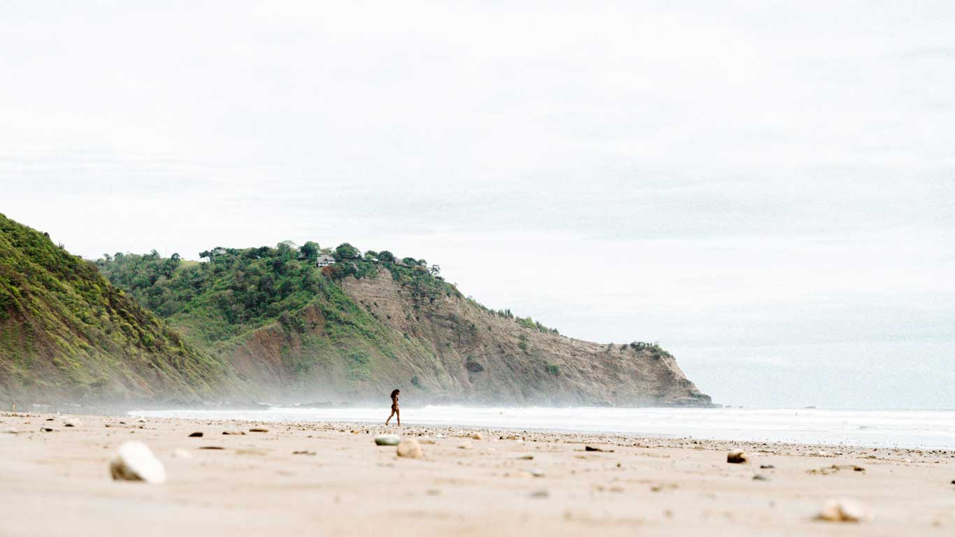 Tranquil landscape of Montañita Beach, Ecuador, with a person walking alone on the sand, surrounded by green cliffs. The sky is overcast, creating a serene and isolated atmosphere.