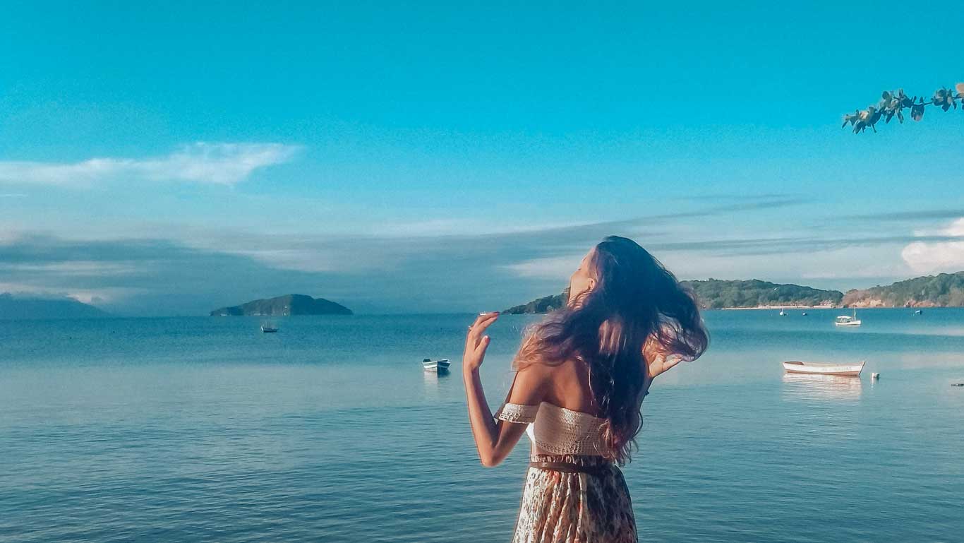 A woman with flowing hair enjoys the peaceful view of Manguinhos Beach in Buzios, Brazil, with gentle waters reflecting the soft sky, boats calmly floating, and distant islands on the horizon, encapsulating a serene tropical escape.