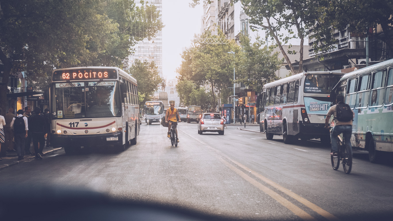 This image captures a typical Montevideo street. Public buses and a cyclist dominate the scene, indicating a common urban commute. The view from the road level adds a dynamic feel, illustrating the everyday motion of life in the city with trees lining the sides, providing a green canopy over the urban environment.