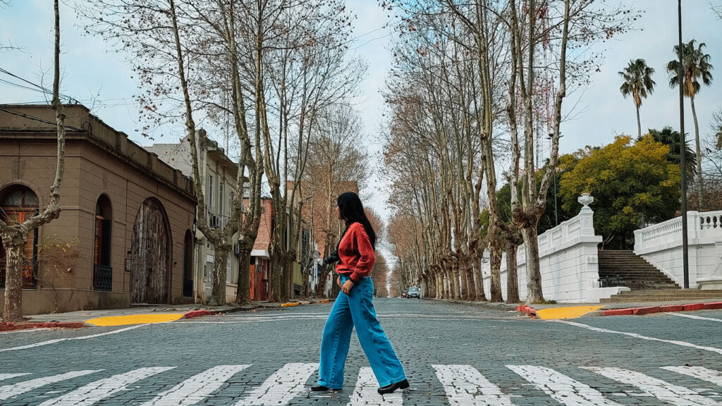 A woman with long hair, wearing blue jeans, boots, and a red hoodie, crossing the pedestrian crosswalk in the old town of Colonia del Sacramento. With colonial houses in the background and a street lined with leafless trees due to winter, fading into the horizon.