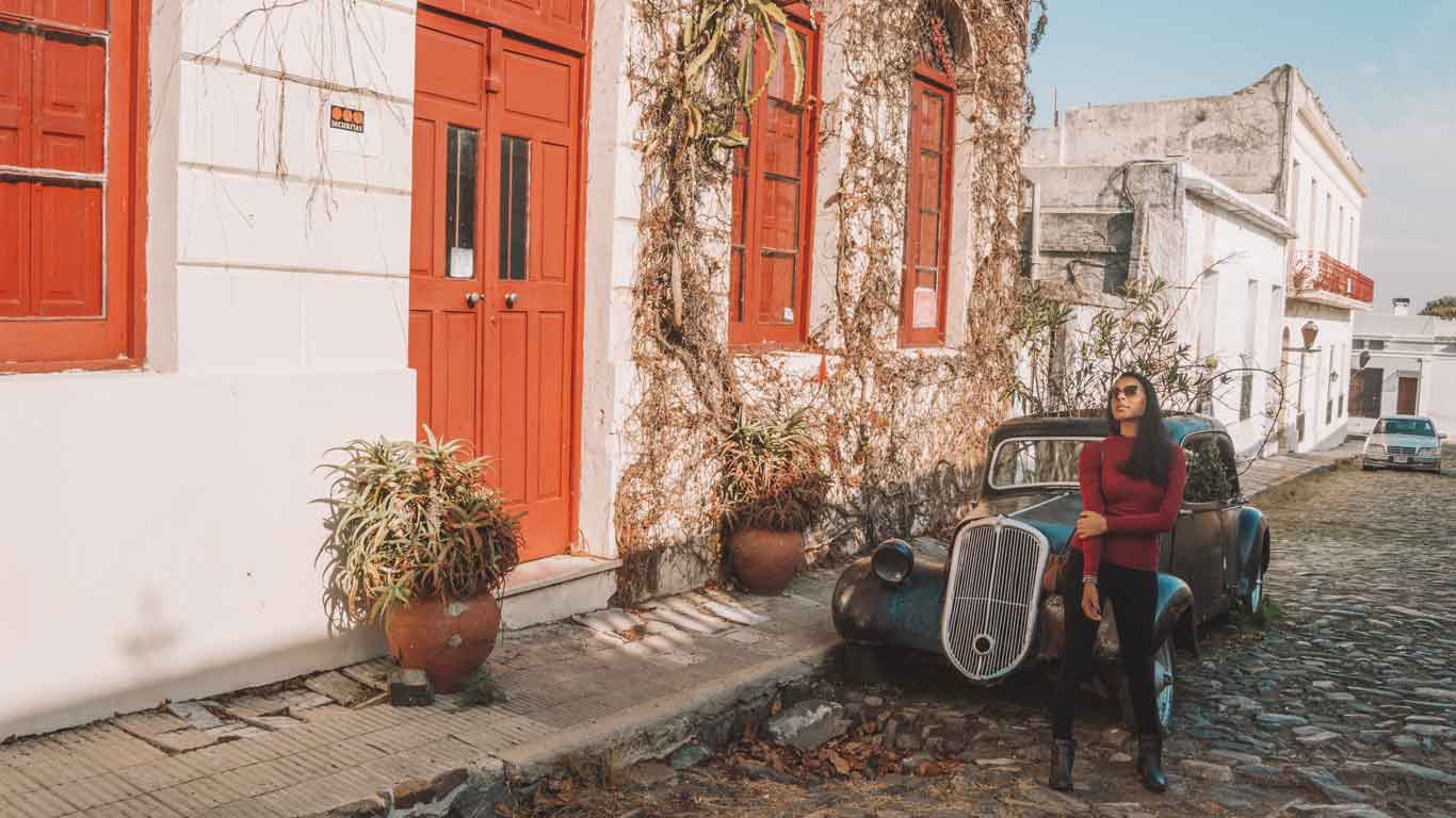 Woman posing next to a vintage car parked in front of a colonial house with colorful doors and windows, typical of the architecture of Colonia del Sacramento.