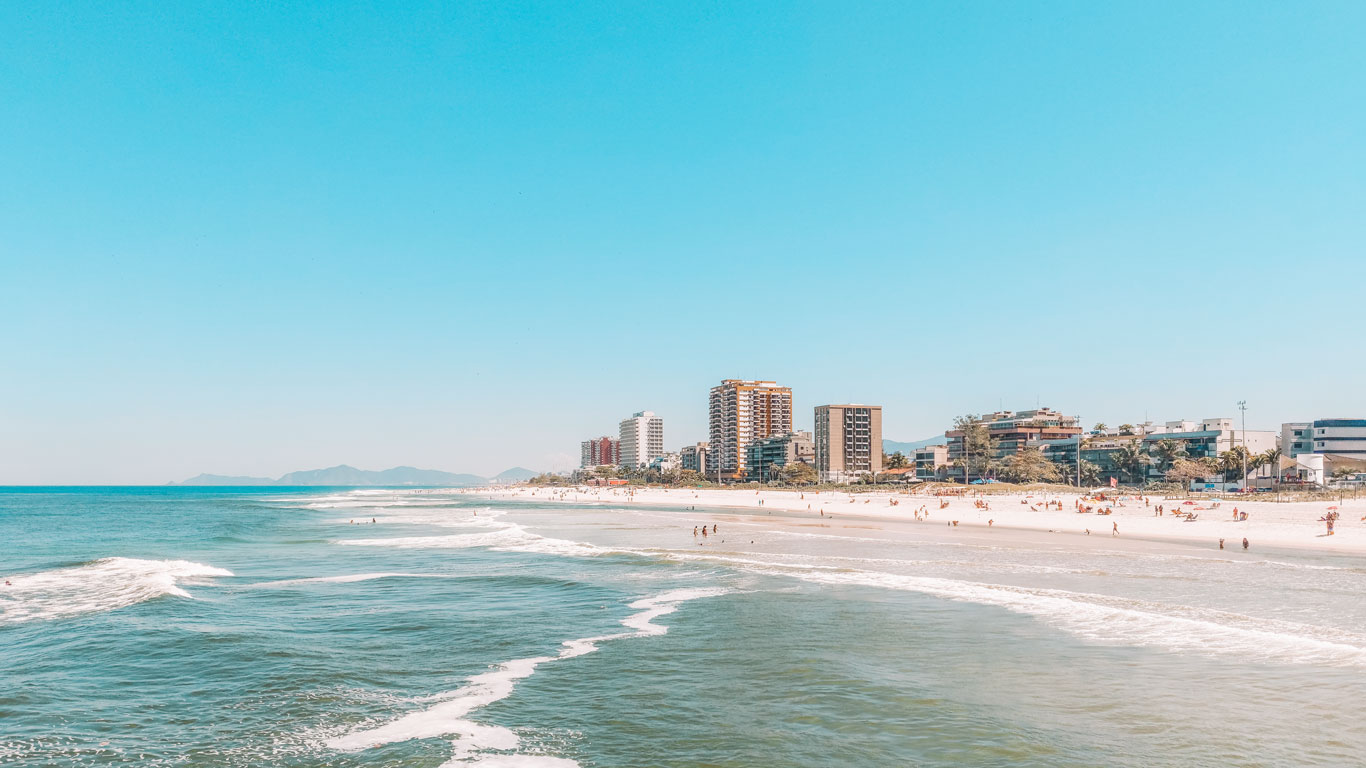 A bustling beach scene at Barra da Tijuca with gentle waves lapping the shore and a backdrop of modern high-rise buildings under a clear blue sky. Beachgoers are scattered across the sandy expanse, enjoying a sunny day in Rio de Janeiro.