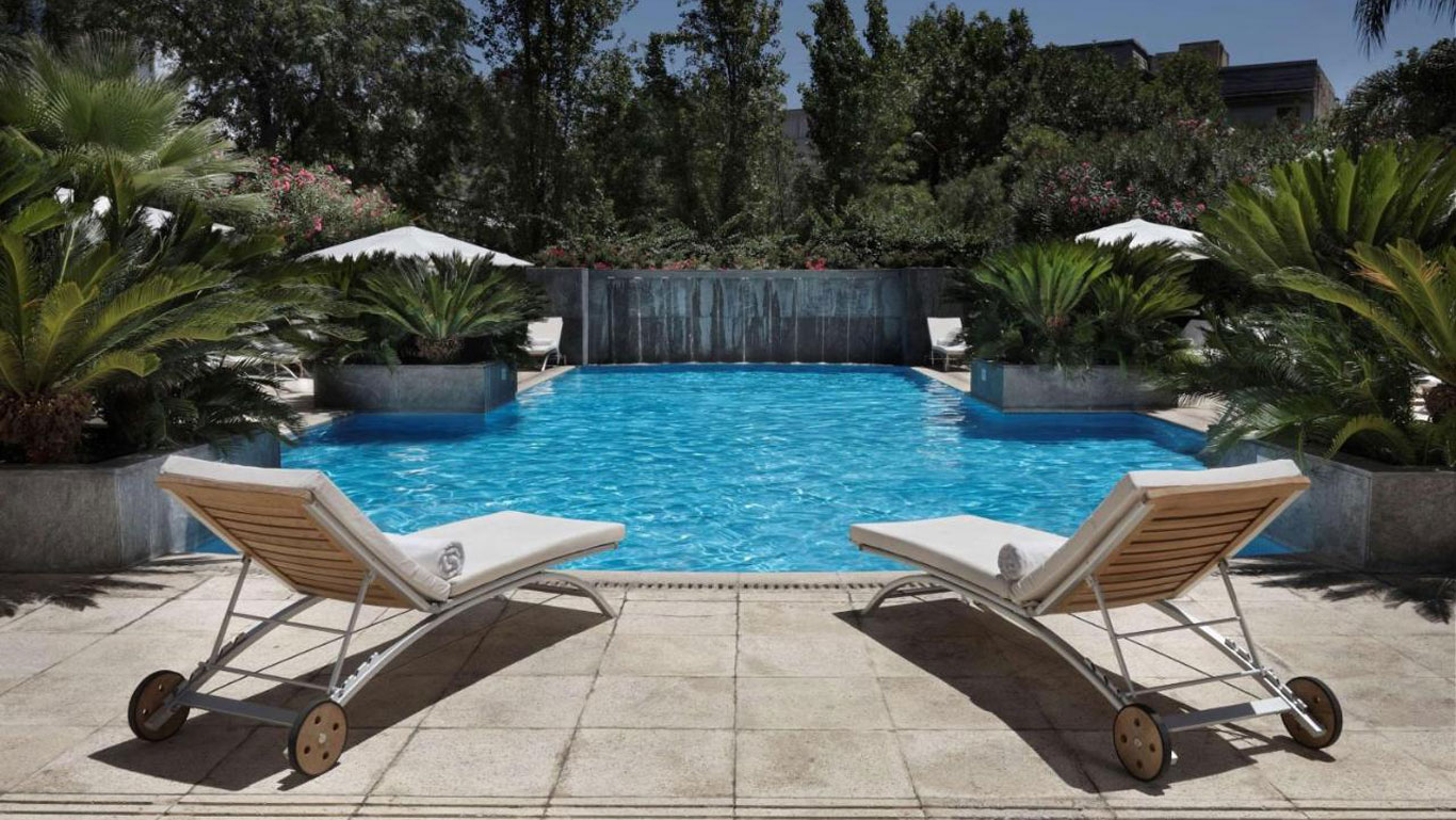 Elegant wooden sun loungers await guests by the serene outdoor pool at Park Hyatt Mendoza, framed by vibrant greenery and a tranquil waterfall feature, offering a taste of luxury and relaxation.