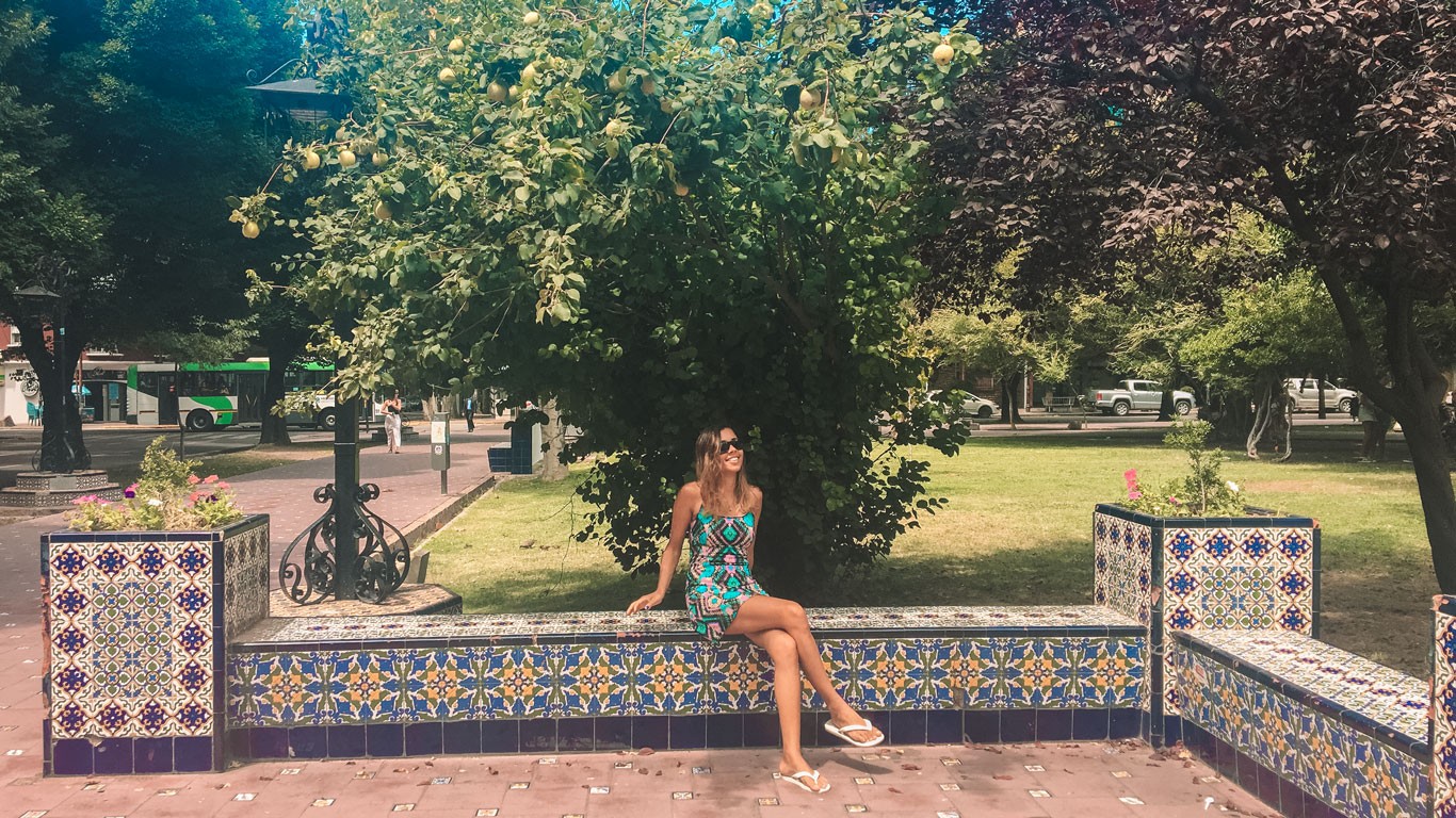 A woman in a green printed dress enjoys a sunny day on a colorful, tile-decorated bench in a park in Mendoza, Argentina, with lush green trees and a vibrant urban backdrop.