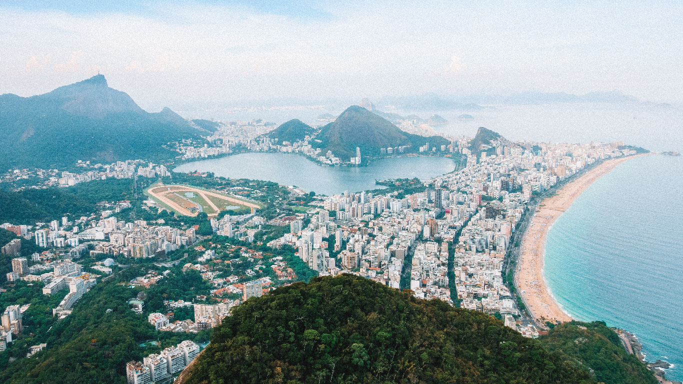 An aerial view of Vidigal favela in Rio de Janeiro reveals a tapestry of densely packed homes cascading down the hillside, with the iconic Rodrigo de Freitas Lagoon and the vast, curving shoreline of Ipanema and Leblon beaches in the foreground. 