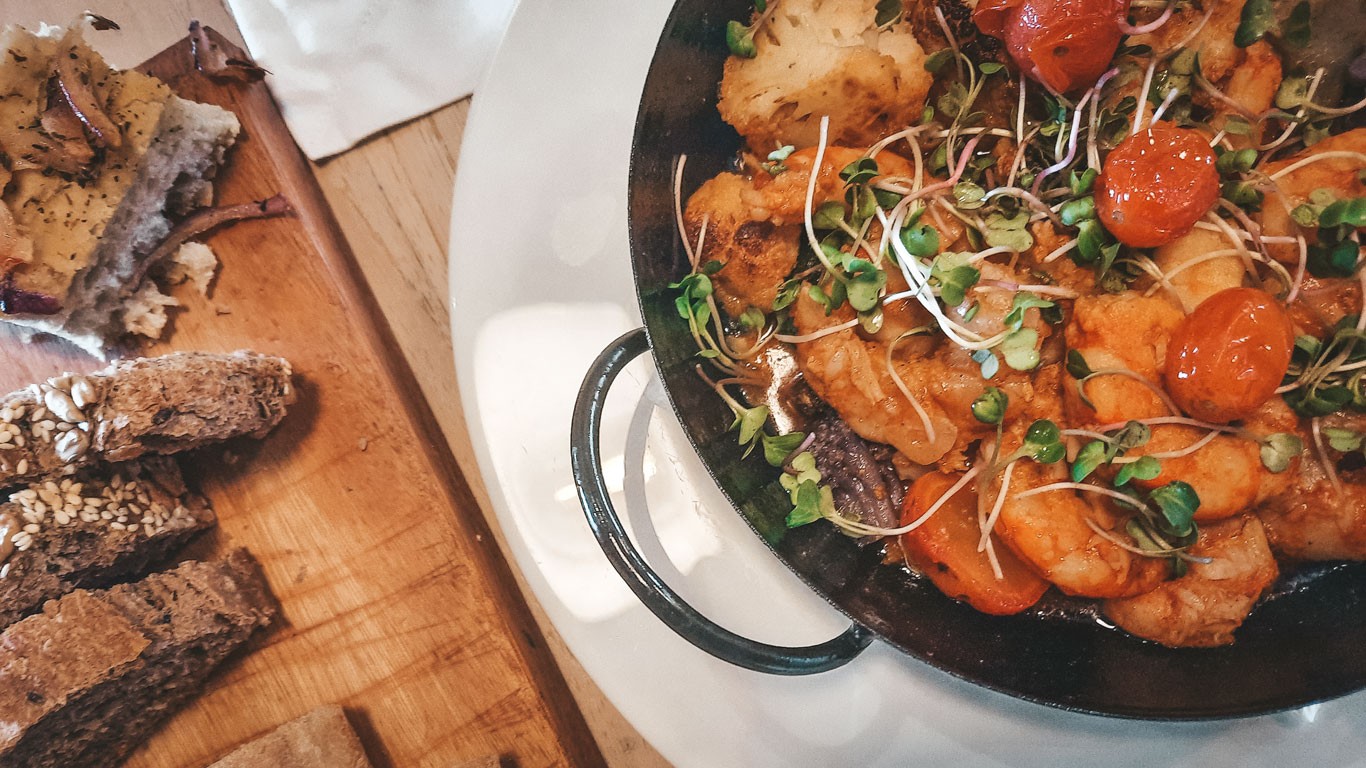 An appetizing presentation of Mendoza cuisine featuring a sizzling pan of golden-browned meats and roasted cherry tomatoes garnished with sprouts, accompanied by crusty whole grain bread on a wooden serving board.