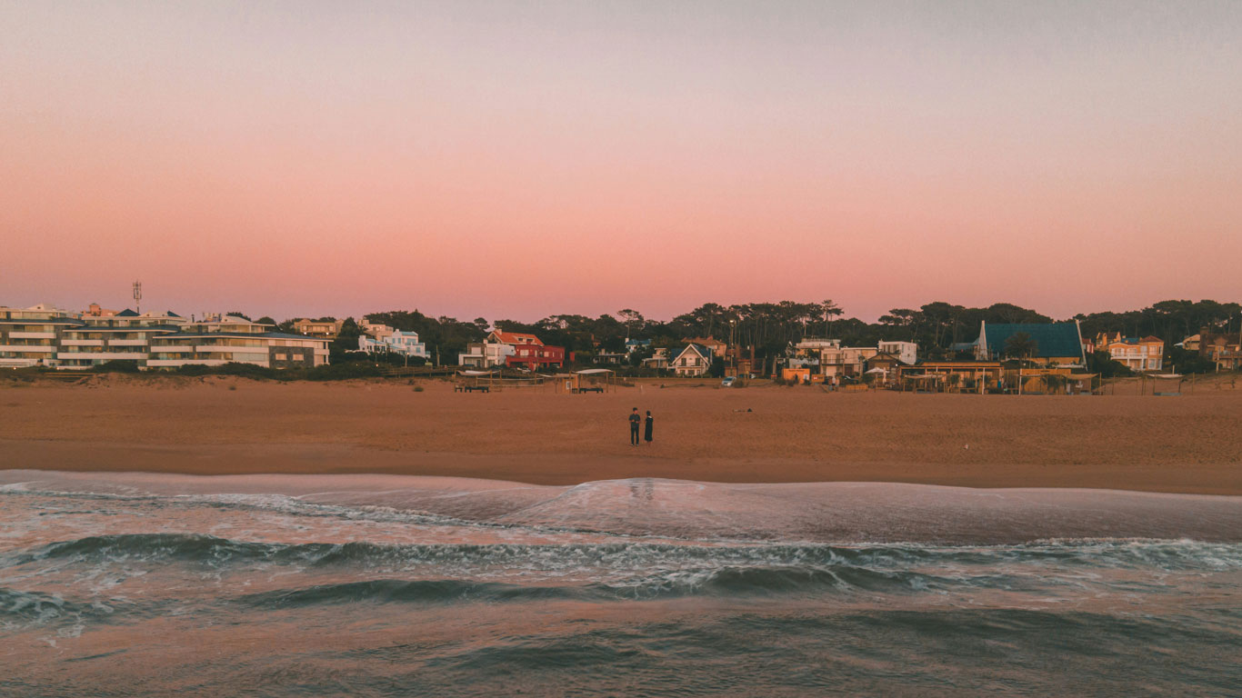 Gentle waves wash onto the sandy shores of La Barra beach in Punta del Este at dusk, with a couple standing near the water's edge and beachfront properties bathed in the soft pink hues of the setting sun."