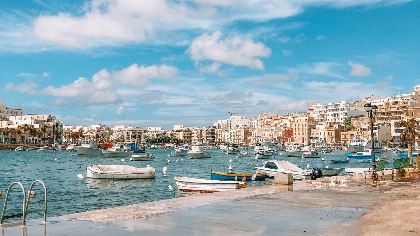 Bright and sunny day in Marsaskala, Malta, with a variety of boats moored in the bay's azure waters, complemented by the picturesque seafront lined with colorful Maltese architecture and leisurely palm trees, evoking a relaxed Mediterranean vibe.