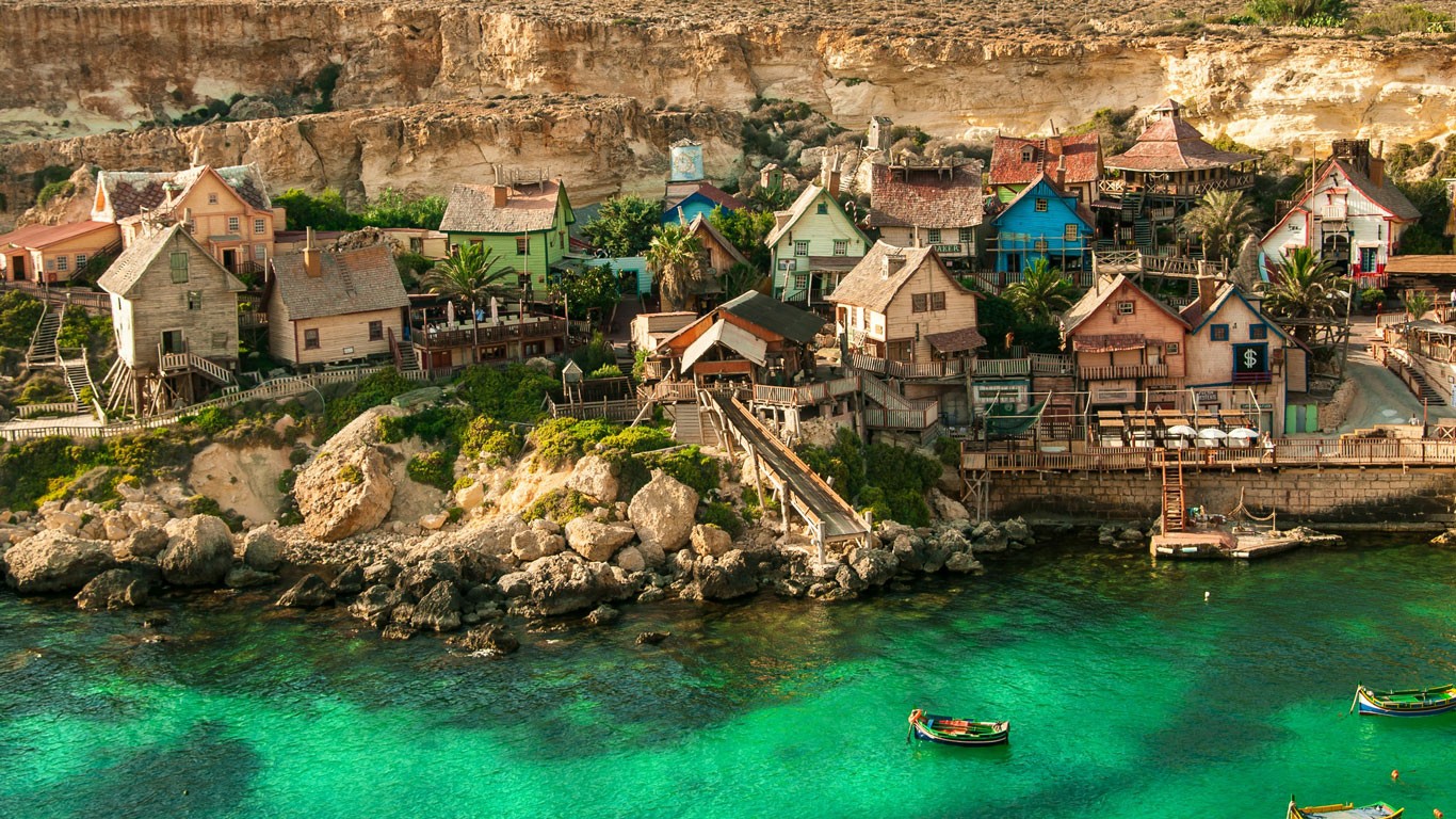 Charming coastal village in Mellieha, Malta, with whimsical structures resembling a movie set, nestled against a rugged cliffside and overlooking clear emerald waters with traditional boats, capturing a fantasy-like Mediterranean atmosphere.