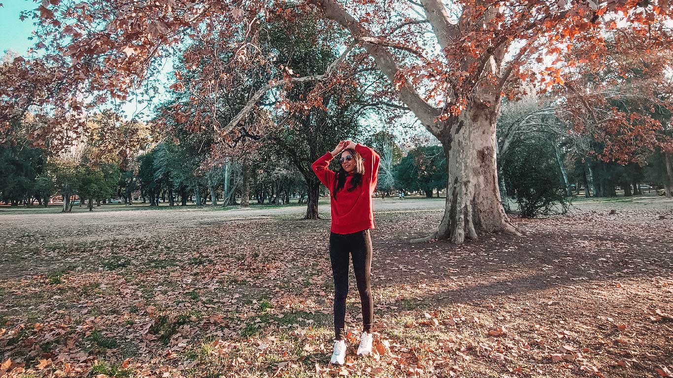 A woman wearing a red sweater and black pants, with her hands resting on her head in a relaxed pose, is under a tree amidst the dry winter leaves in Parque General San Martín in Mendoza.