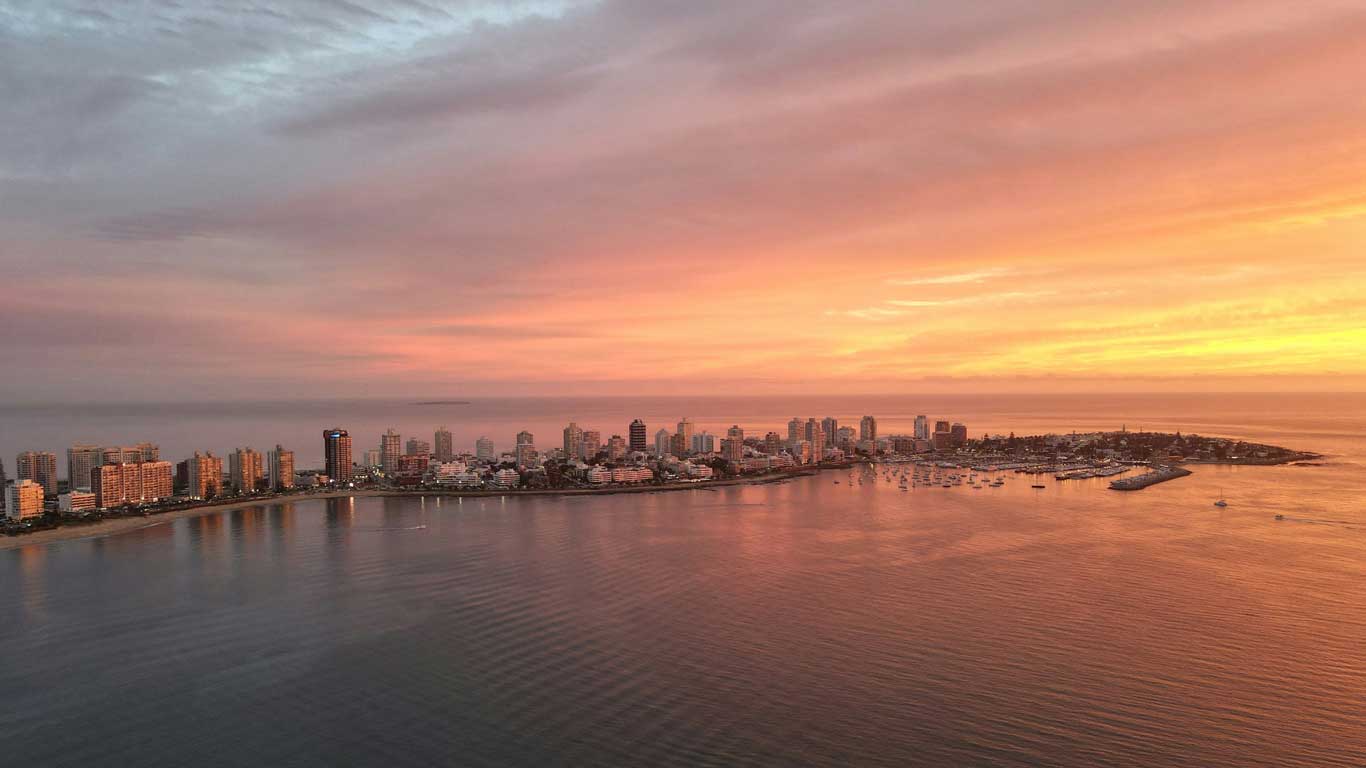 Aerial view of Punta del Este, Uruguay, at sunset with the skyline silhouetted against a pastel-colored sky, reflecting over the calm sea waters.