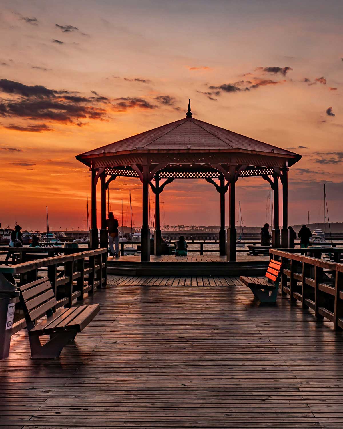 Twilight at the peninsula in Punta del Este, featuring a wooden pier leading to a gazebo with silhouetted figures enjoying the view, against a backdrop of moored sailboats under a vibrant orange sky.