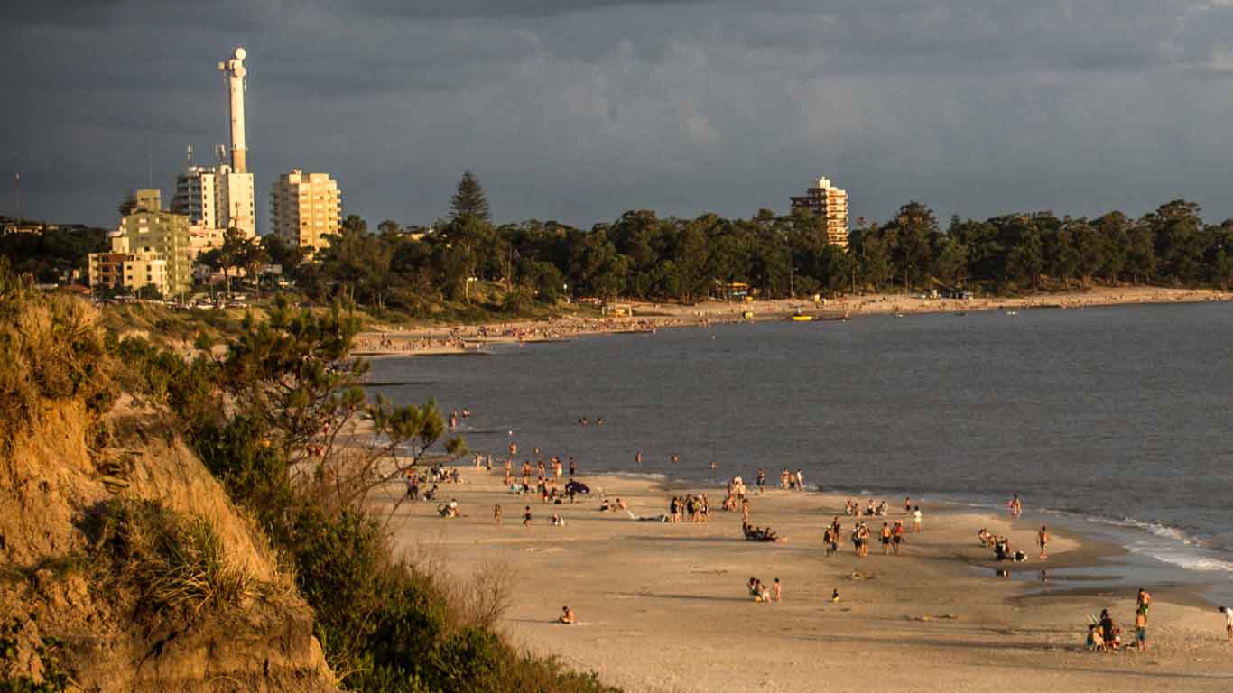 A scenic view of Piriápolis in Punta del Este, featuring a sandy beach with numerous people enjoying the water and shoreline. In the background, there are buildings and a lighthouse set against a backdrop of lush trees under a cloudy sky.