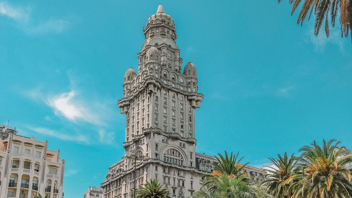 The image features Palacio Salvo, an iconic landmark in Montevideo, Uruguay. This historic building features complex architecture with ornate details and a commanding presence against a clear blue sky, surrounded by palm trees and adjacent buildings. He can be known on the best tours in Montevideo.