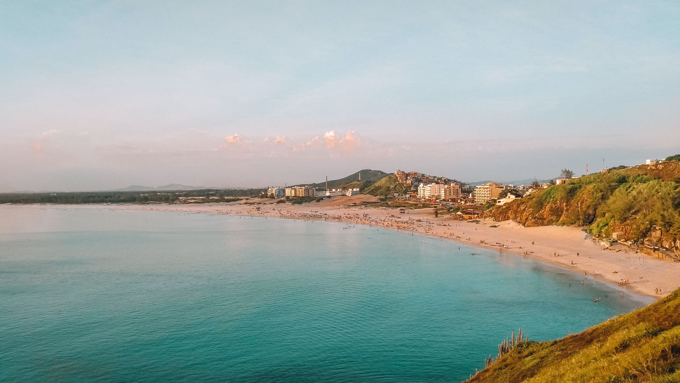 An expansive view of Grande Beach in Arraial do Cabo, captured during a serene sunset. The beach stretches across the image with scattered visitors enjoying the vast sandy shores. The calm turquoise waters contrast beautifully with the soft pastel sky, while the urban landscape hugs the coastline, creating a picturesque scene that blends natural beauty with city life.