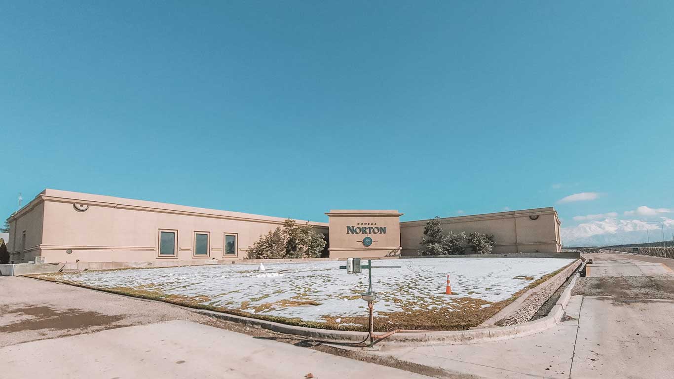 Exterior view of Bodega Norton in Mendoza, Argentina. The large beige building with the winery's name prominently displayed on a central wall sits under a clear blue sky. Snow dusts the surrounding ground and distant mountain peaks, adding to the serene winter landscape.