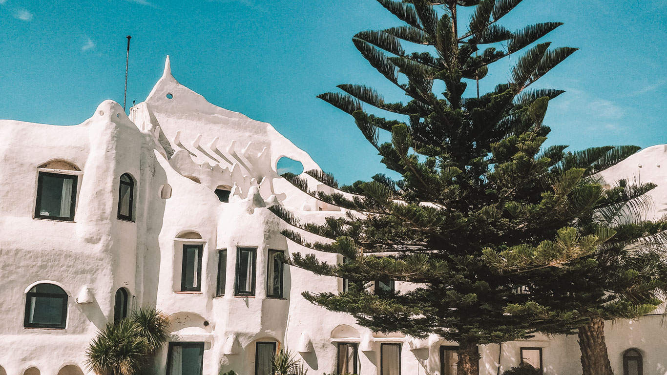 This image shows the unique architecture of Casa Pueblo in Punta Ballena, Uruguay. The white-washed, whimsical building features irregular shapes and organic forms, complemented by a tall, lush tree in the foreground under a clear blue sky.