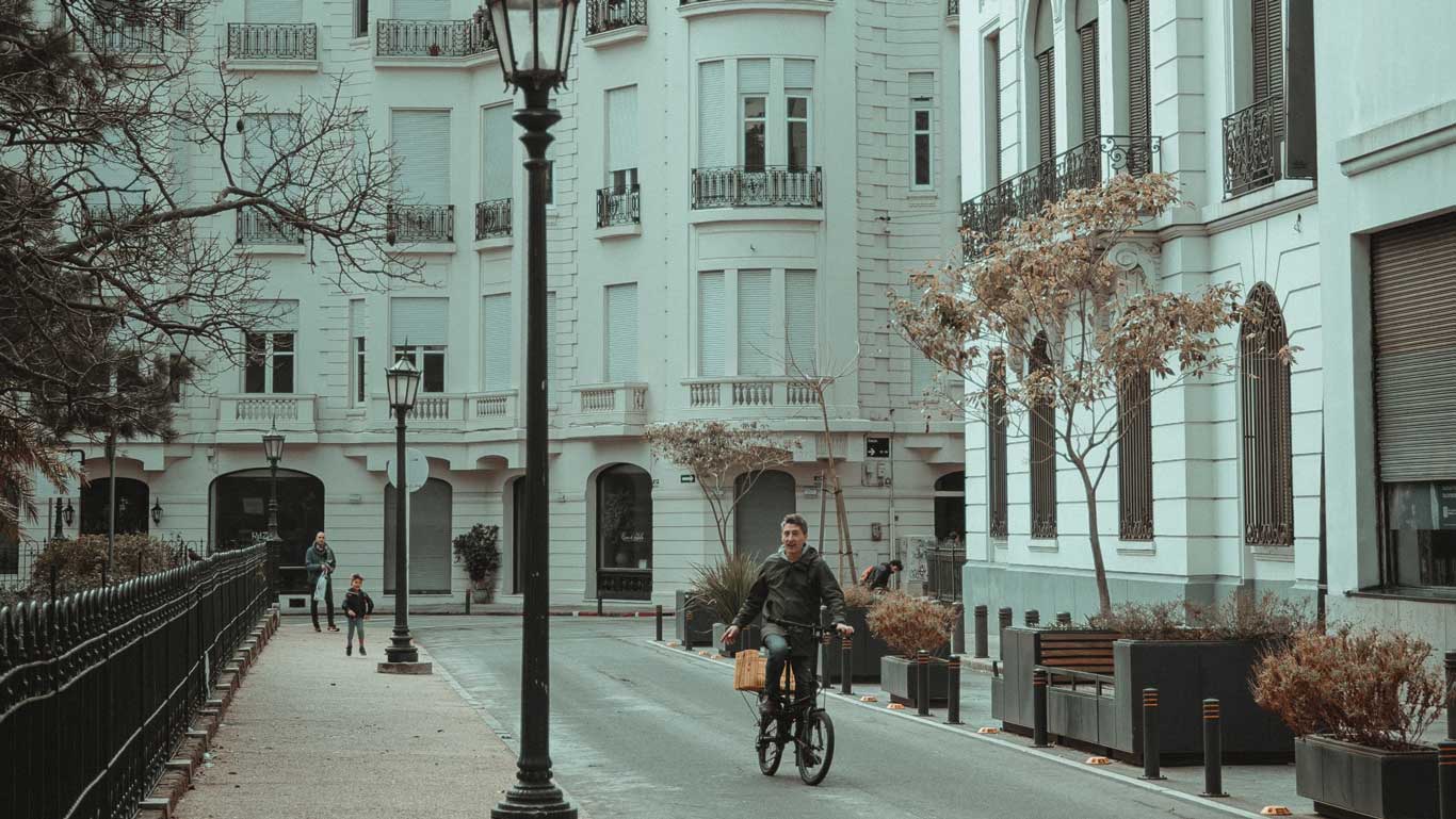 The image depicts a charming street scene in Montevideo, Uruguay. A man rides a bicycle along a quiet street lined with elegant, white buildings featuring ornate balconies. In the background, a parent and child are walking, adding to the serene and picturesque urban atmosphere.