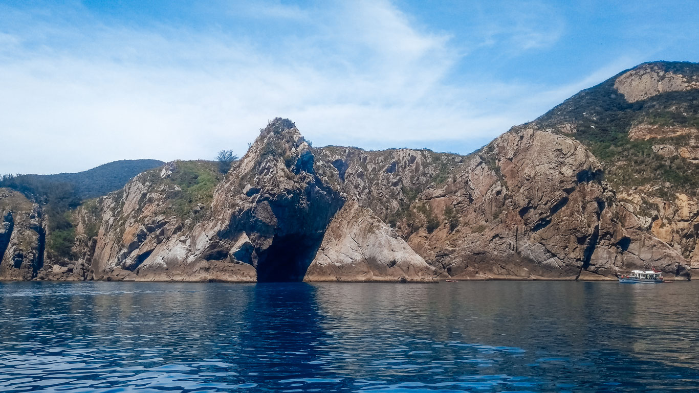 The image showcases the breathtaking Gruta Azul in Arraial do Cabo, with its distinctive rocky cliffs forming a natural archway over the azure waters. The towering cliffs, jagged and covered in sparse vegetation, reflect off the calm sea, creating a serene and majestic landscape. A small boat is visible in the distance, giving scale to the grandeur of the rock formations, emphasizing the popular attraction for visitors looking to explore the natural wonders of this area.