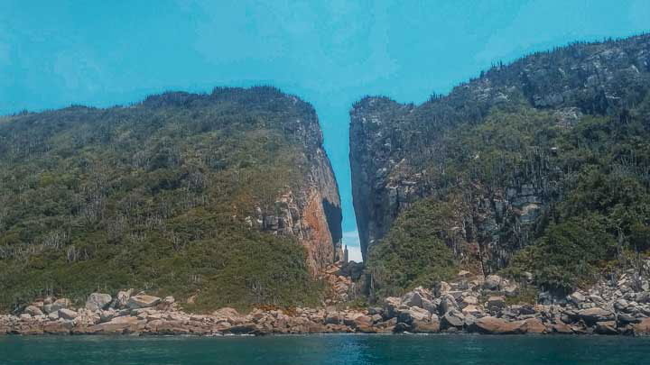 This image depicts the stunning natural formation known as Fenda da Nossa Senhora in Arraial do Cabo. The view shows a large, narrow split between two rocky cliffs covered with dense vegetation, leading to a hidden cove. The turquoise waters in front and the rugged, untouched landscape offer a captivating scene that highlights the unique geological features of this coastal area.