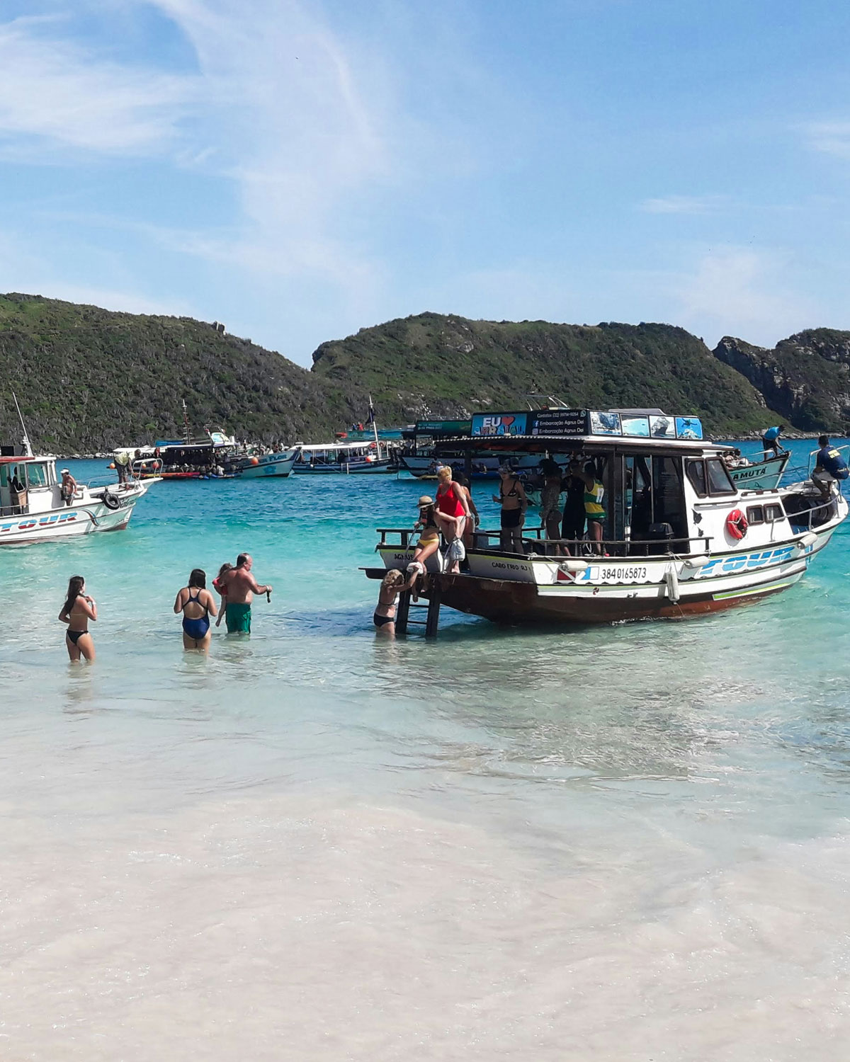 Tourists enjoy the clear turquoise waters at Farol Beach in Arraial do Cabo, Brazil. Multiple boats are moored near the shore, with people disembarking into the shallow water. The scene captures a bustling day at one of the most popular beaches in Arraial do Cabo, surrounded by lush green hills under a bright blue sky.