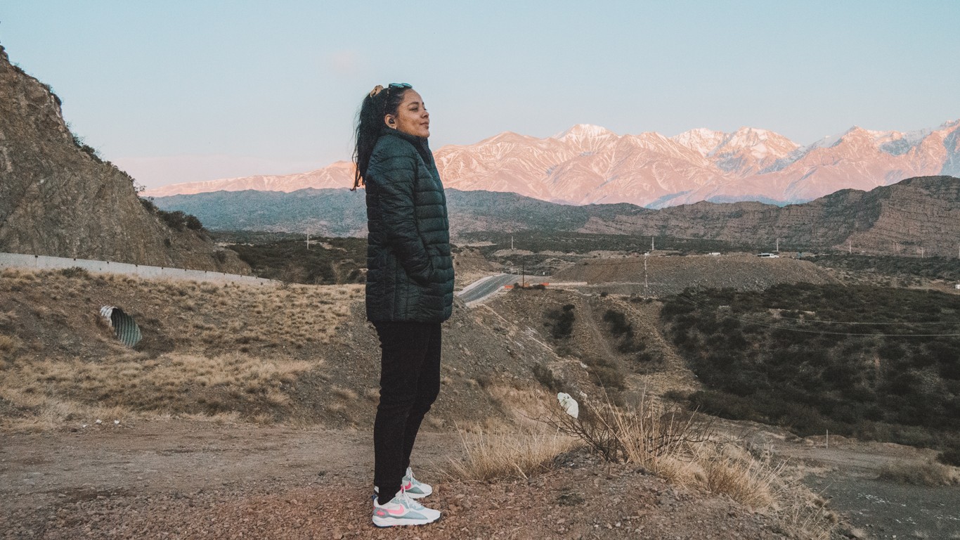 A woman stands on a dirt path with her hands in her pockets, wearing a black jacket and sneakers, gazing into the distance. Behind her, rugged terrain stretches out to snow-capped mountains under a clear sky, capturing the serene landscape of Mendoza in August.