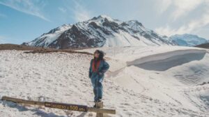 A person dressed in winter gear stands confidently on a wooden post, surrounded by snow in Mendoza, Argentina. Behind them, towering snow-capped mountains rise against a clear blue sky, with a sign below that reads "INGRESO a 500 mts."