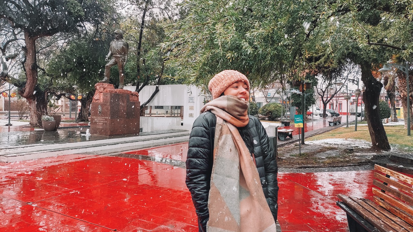 A woman dressed warmly in a black jacket, scarf, and beanie stands smiling in a snowy park in Mendoza. Behind her is a statue on a red stone pedestal, surrounded by trees and a few benches, with snowflakes falling gently.